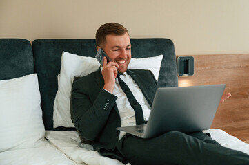 Smiling, lying down on bed and talking by phone. Businessman is indoors in the hotel room