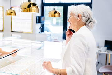 Portrait of grey haired senior female consultant, employee in gewelry store, shop. Stands with a tablet, talking smart phone, consulting. In elegant clothes.