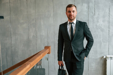 Beautiful man in suit and tie is standing indoors and holding case in hand