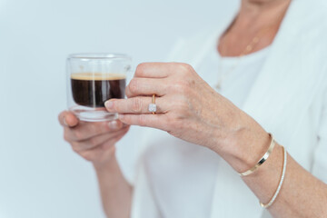 Close up of grey haired senior woman drinking coffee show diamond ring and gold bracelets on white background.