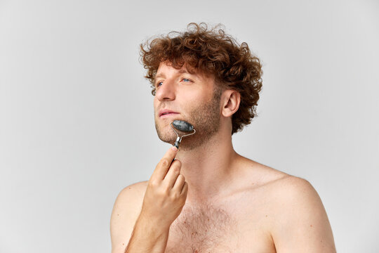 Portrait of matue man with curly hair, standign shirtless and doign face massage with roller against grey studio background. Concept of mens beauty, body and skin care, cosmetology, wellness, ad