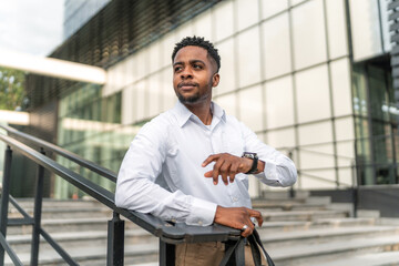 A portrait of a handsome Black businessman leaning against a railing during his break, holding a...