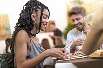 African-American woman with afro hairstyle slices pizza at Italian pizzeria with Caucasian friends....