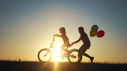 Foto op Canvas mom teaches daughter to ride a bike. happy family childhood dream concept. mom and little daughter learn to ride a bike silhouette in park in nature. happy family sunlight goes in for sports outdoors © maxximmm