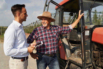 Senior farmer and sales representative next to tractor talking over digital tablet
