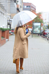 Naklejka premium A girl in a beige coat with a transparent umbrella walking down the street. Europe. Travels. autumn Coldly. Weather forecast.