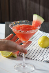Hand picking a  margarita served in a martini glass. Lime and watermelon slices rest on a black and white striped cloth sitting on a white marmbled surface, with harsh daylight coming from behind.