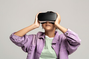 Teenager girl in virtual glasses goggles watching movie playing video games on gray background. Smiling teen enjoying virtual world in VR helmet. Cyberspace, entertainment, online recreation concept.
