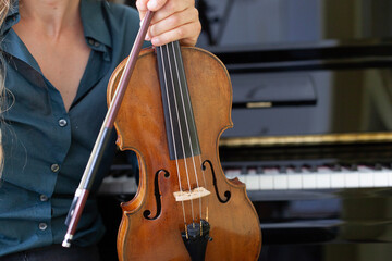 Frau hält Geige in Hand mit Klavier im Hintergrund.
Woman holding violin in hand with piano in background.