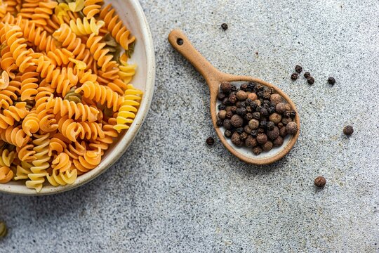 Overhead view of a bowl of uncooked fusilli pasta and a spoonful of black peppercorns on a table
