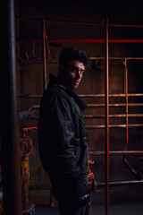 despaired unshaven man looking at camera next to rusty pipelines in abandoned post-disaster subway