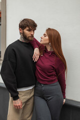 Pretty young urban stylish couple in fashion casual clothes with a hoodie are standing together on the street. Handsome hipster man and beautiful red-haired woman