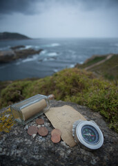 A traveler's compass, an old sheet of paper, a glass bottle with a message inside and several metal coins lie on a rock against the backdrop of a sea bay - 647611610