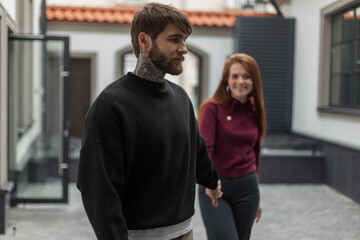 Cool handsome brutal hipster man with a beard and tattoo in a black pullover and a beautiful happy redhead girl with a smile in a hoodie on the street. Beautiful fashion couple walks hand in hand