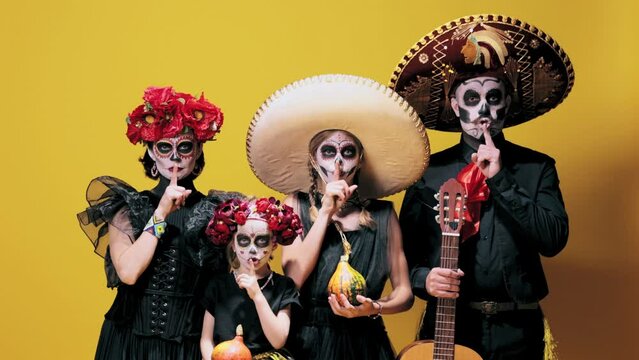Shhh. Keep silence. Man, woman and daughters in sugar skull makeup and costumes over yellow background. Concept of halloween, El Dia de Muertos, holidays and festivals, mexican traditions, family
