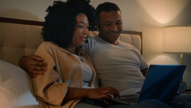 Happy family man and woman girlfriend and boyfriend African American couple in bed night home evening shopping with laptop together choosing goods online internet buying talking discuss using computer