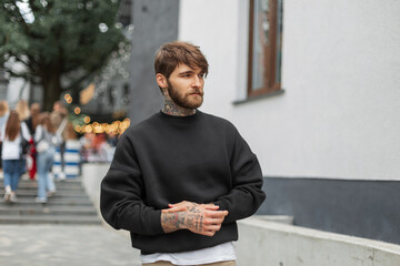 Cool handsome brutal hipster man with a beard and tattoo in a fashion black mockup sweatshirt walks...