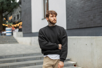Cool handsome young hipster man with hairstyle and beard in a black fashionable sweatshirt walking...