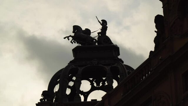 powerful cinematic scene of a roman warrior riding a chariot with two horses raised arm holding a sword in the sky sculpture on top of a roof silhouette dramatic clouds moving quick speed background