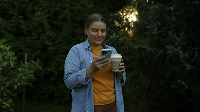 Attractive young blonde businesswoman holding takeaway coffee cup, using mobile phone outside in city park at sunset. High quality FullHD footage