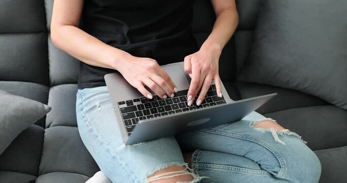Woman in torn jeans sitting on black sofa with laptop in living room. Contemporary concept of social networking and remote work slow motion