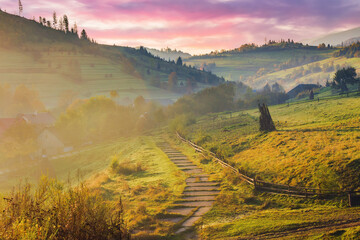 mountainous countryside on an autumn foggy weather at sunrise. empty rural fields on the hills. path to the village. mountain ridge in the distance beneath a sky with purple clouds