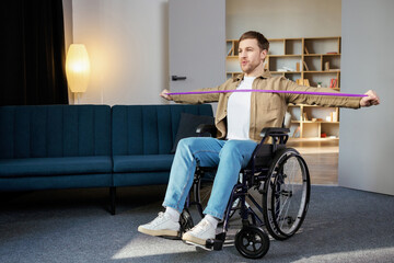 Young disabled man in wheelchair doing exercises with rubber band at home. Caucasian handicapped guy working out in living room