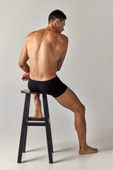 Back view image of young man with muscular body sitting on chair shirtless in underwear against grey studio background. Concept of men's health and beauty, body care, fitness, wellness, ad