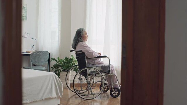 Long shot of depressed elderly woman with disability in wheelchair looking at window in nursing home