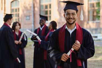 Portrait of happy student in graduation gown smiling at camera while standing with diploma