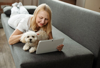 Happy mature woman lying on sofa, using tablet and internet app for videocall while pet dog nearby. Concept of using gadgets and technology by older people