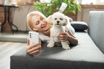 Happy senior woman lying on sofa, holding phone in hand and using internet app for videocall while pet dog nearby. Concept of using gadgets and technology by older people