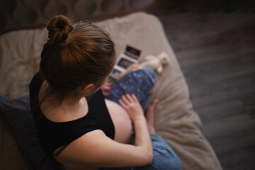 beautiful young pregnant woman sits on the bed in the bedroom and holds her baby's tiny clothes in her hands