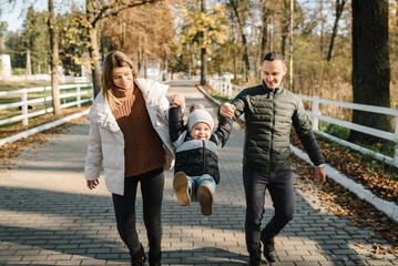 Mother, father throws up kid and running in street near forest in nature. Mom, dad hold hands son child walking in park at sunset. Family spending time together on vacation. Concept of autumn holiday.
