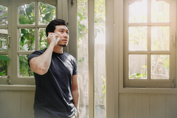 Fototapeta premium Asian man is talking on phone standing next to window.