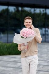 Fototapeta premium Flower delivery. Smiling delivery man with luxury bouquet of hydrangea flowers talks on smartphone with client on territory modern residential building. Gift for Valentines day, Birthday, Anniversary