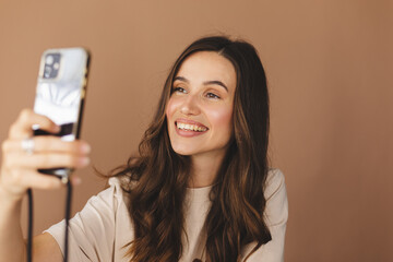 Young smiling brunette curly woman wear beige t-shirt doing selfie shot on mobile phone post photo on social network isolated on beige background. Woman make video call, selfie on her mobile phone.