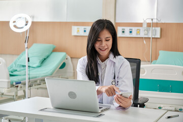 Obraz na płótnie Canvas Young beautiful Asian female doctor online working with laptop and smartphone on medical consultation application via internet at working desk in hospital clinic.