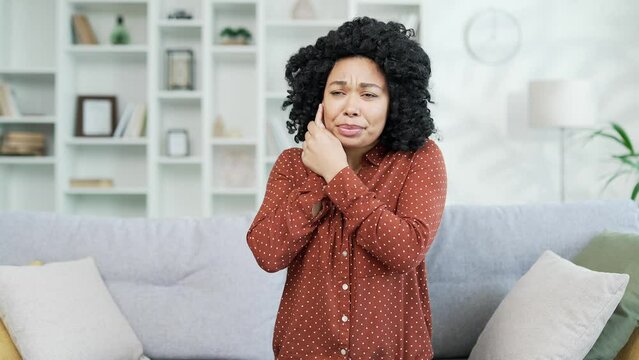 Earache. Sick african american female suffers from ear pain sitting on sofa at home. Upset black woman touches her ear with her fingers. She has furuncle, shingles, eczema, ear diseases, otitis media