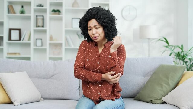 Young african american female in a shirt suffering from elbow pain while sitting on the sofa in the living room at home. Sad black womant is massaging a joint, rubs her hand, feels discomfort in joint