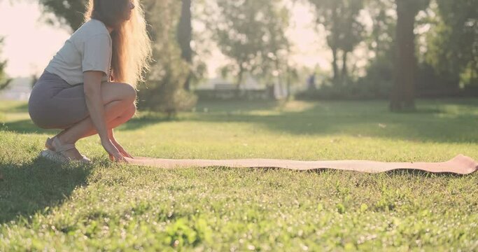 Woman lay out yoga mat on lawn in park. Side view, sunshine, morning, sunrise, green grass