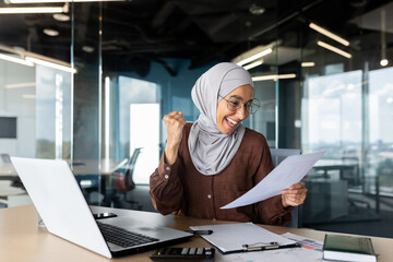 Happy young muslim business woman in hijab is happy with success and achievement, sitting at office table and holding documents, deal, financial results.