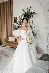 Bride. Young fashion model with perfect skin and makeup in room. Luxury wedding day. Fashion photo. A charming bride in a stylish dress holds a bouquet of white roses and gypsophila flowers at home.