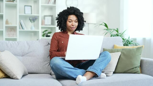 Young african american female freelancer typing on laptop sitting on sofa at home. Black woman student works on a computer, chats online with a friend, writes an email message or browses social media