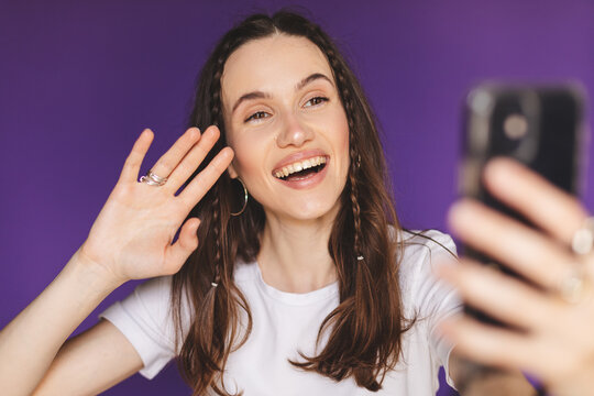 Happy young woman with two thin braids blogger influencer holding smart phone wave hand hello. Smiling vlogger girl looking at mobile make video call, shooting vlog taking selfie on purple background.
