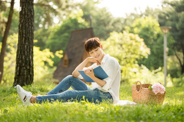 A young man is having a good time sitting in a forested park reading a book.