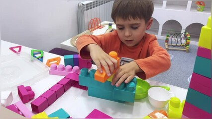 Little toddler boy playing with toys sitting on the chair among scattered toys, a mess in the children's room. He loves to assemble a constructor. 4k footage