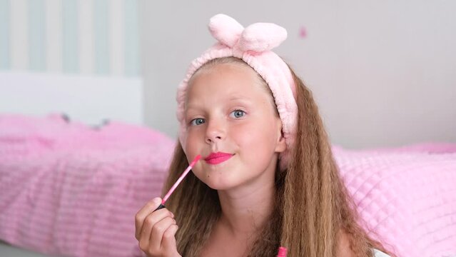 A girl sits near the bed and paints her lips. The child looks at the camera and applies lip balm.