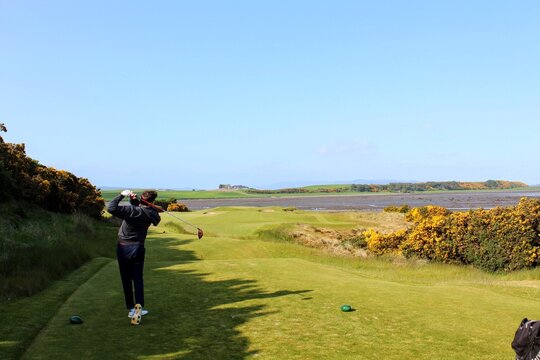 A young man swinging a golf club on a tee box surrounded by the beautiful views of the sea in the background, outside Inverness, Scotland