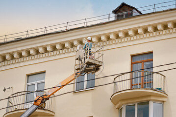 Fototapeta na wymiar Man paint wall with spray gun on aerial lifting platform. Painter working in crane bucket, paint building wall of historic building with paint spray gun. Repair and restoration facade of building